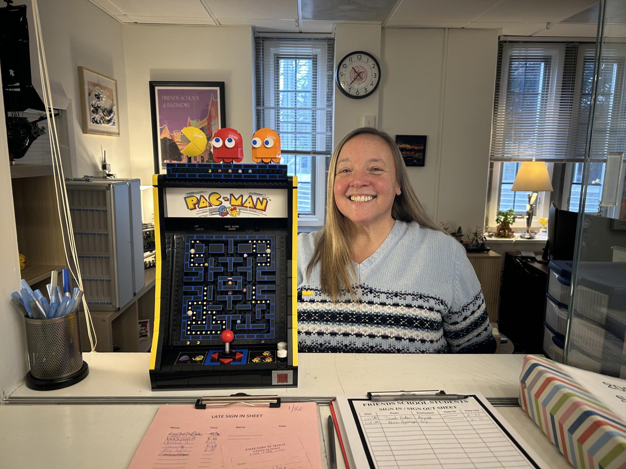 Ms. Wilmoth smiles in the front office window, next to a desktop Pac-Man game she built out of Legos. More of her Lego projects, from Katsushika Hokusai's "The Great Wave off Kanagawa" to cherry blossom trees, are visible behind her.